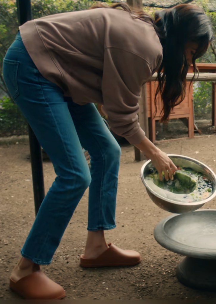 Meghan Markle wearing brown clogs while bending over to place food into a bird bath.