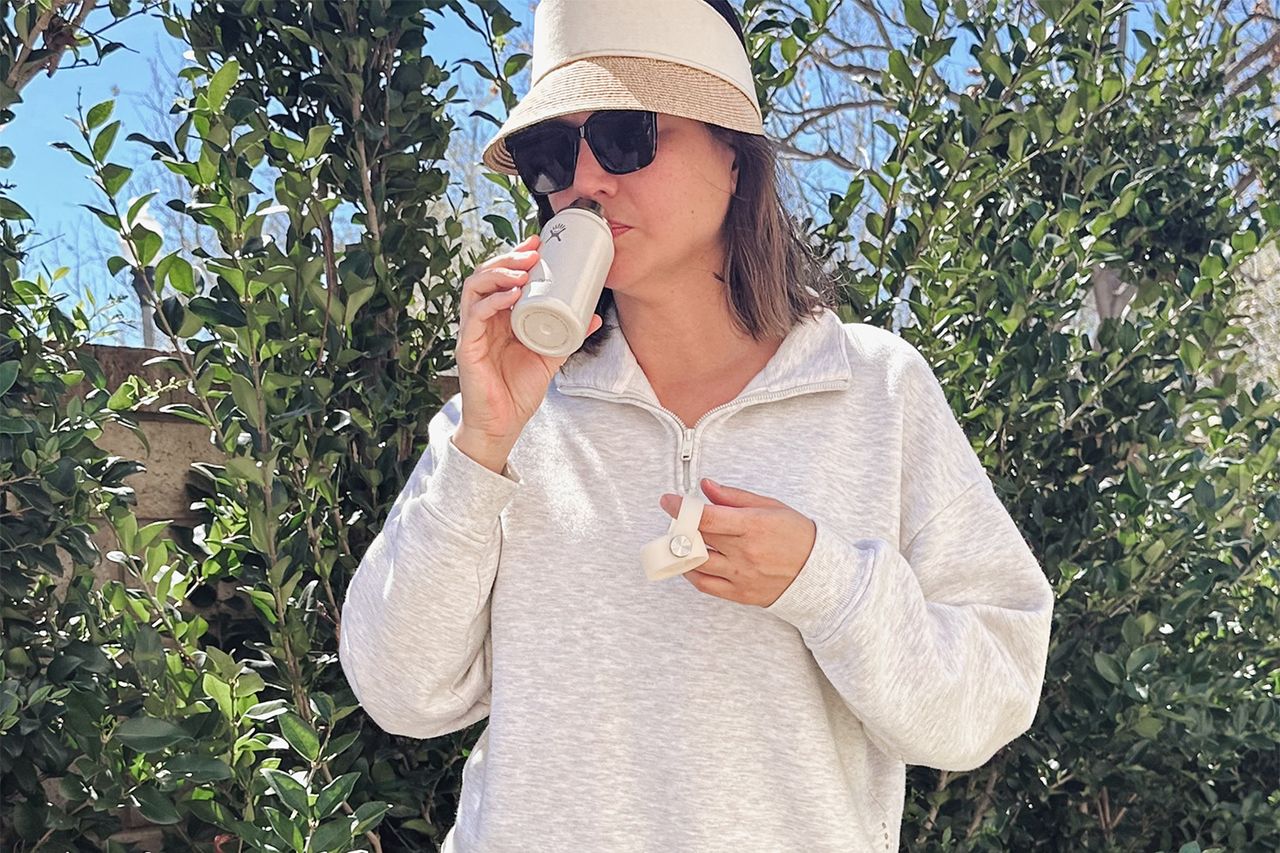 A woman stands outdoors wearing a hat and sunglasses sipping from a small drink container surrounded by green foliage Credit: People