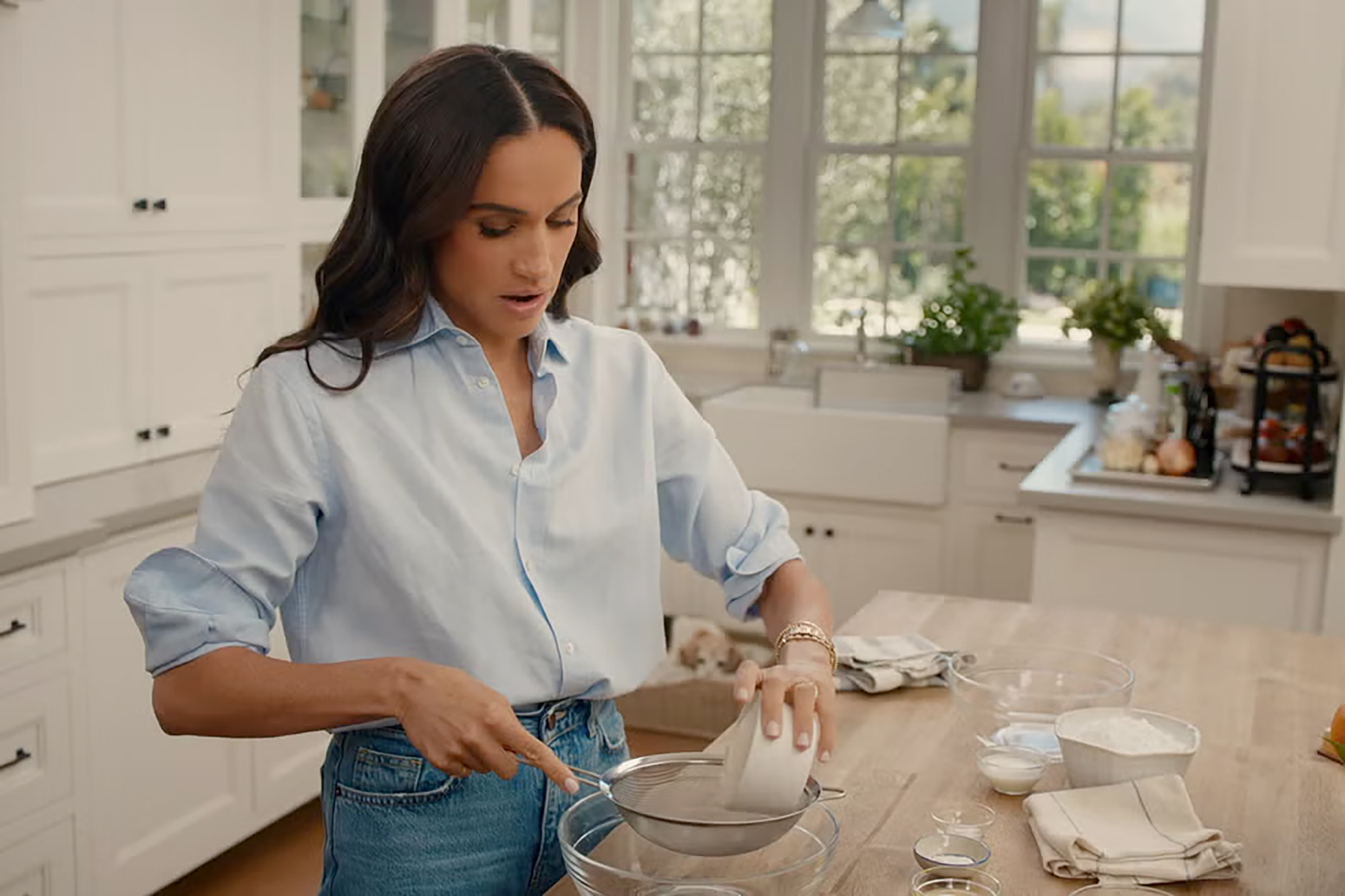 Meghan Markle sifting ingredients in a light blue shirt in a kitchen.