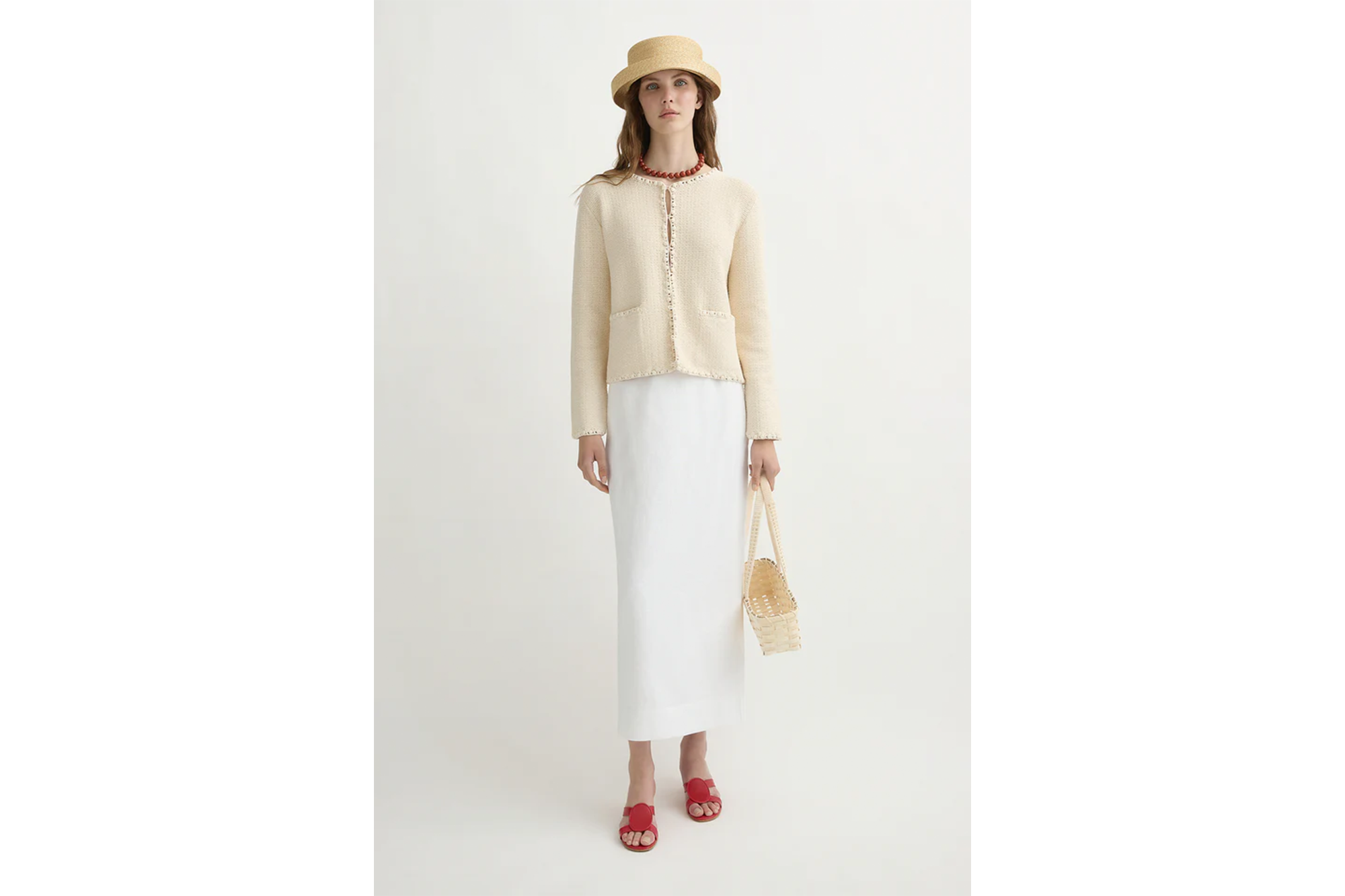 A woman models an off-white jacket with a white skirt, red sandals, and a straw hat and bag.