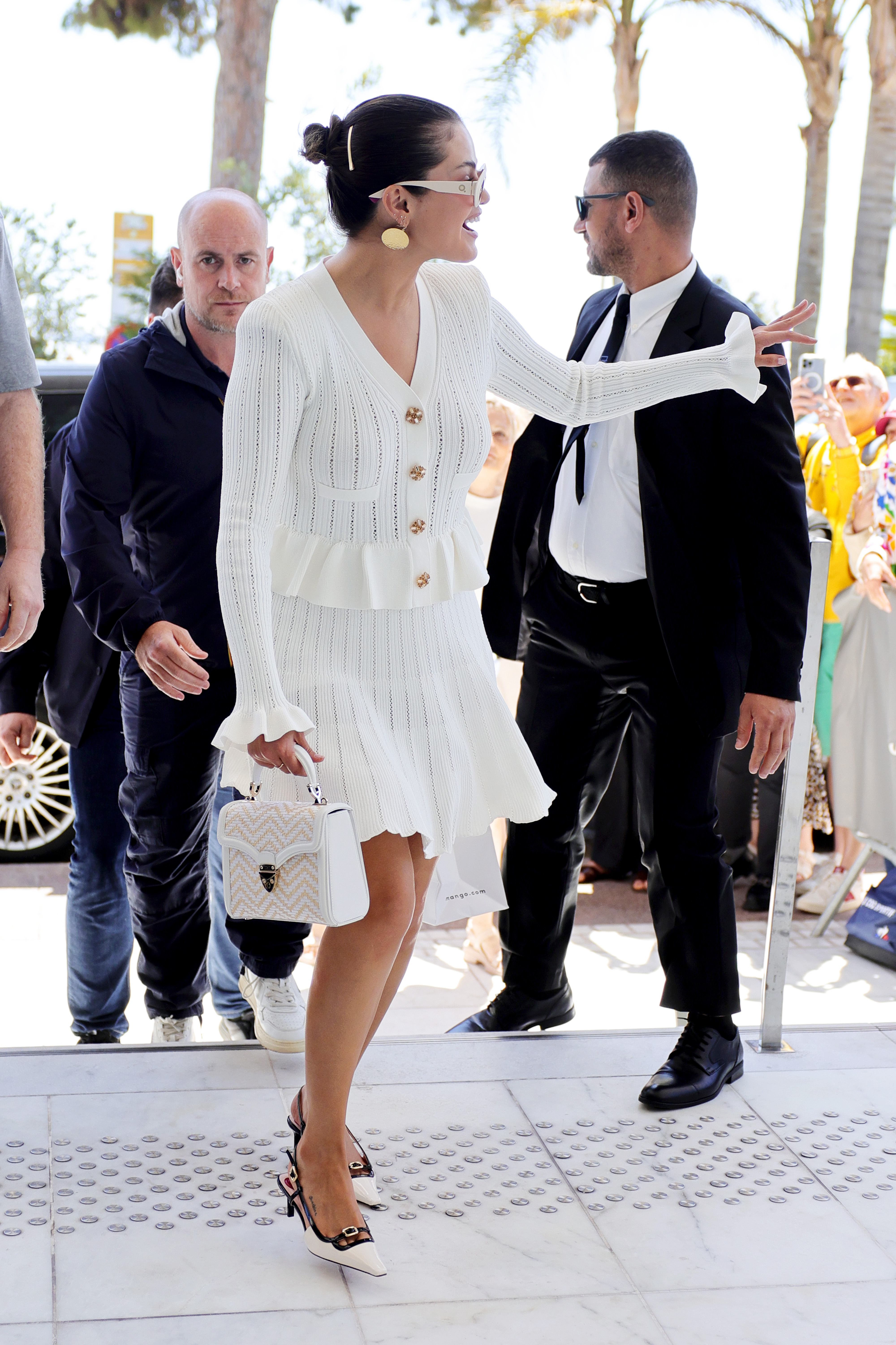 Selena Gomez in a white knit outfit with a peplum top and a matching skirt, holding a white patterned handbag, at the Cannes Film Festival.