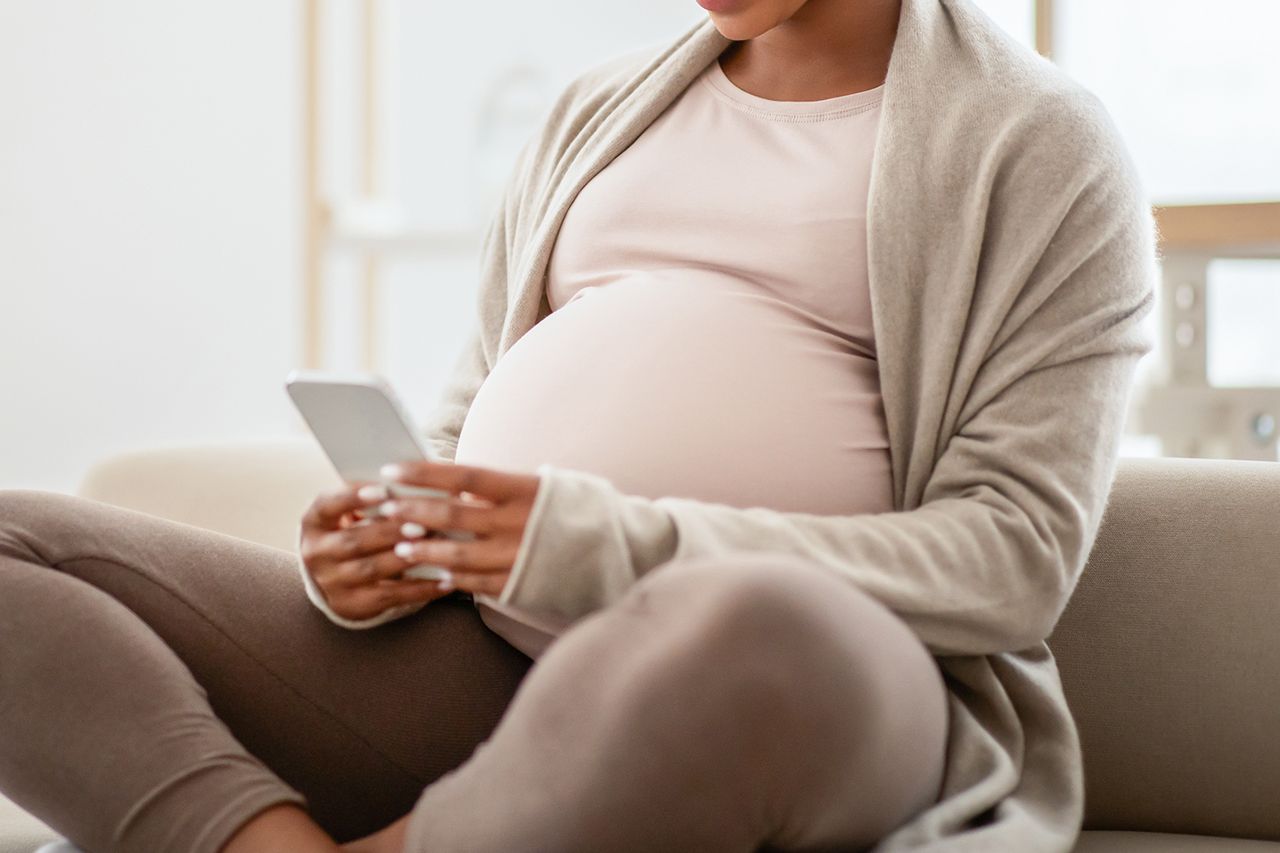 Beautiful young pregnant woman watching funny video on smartphone, using mobile while sitting on couch at home. Millennial expecting african american lady using cellphone, mobile app, copy space Stock image of a pregnant woman looking at her phoneCredit: Getty