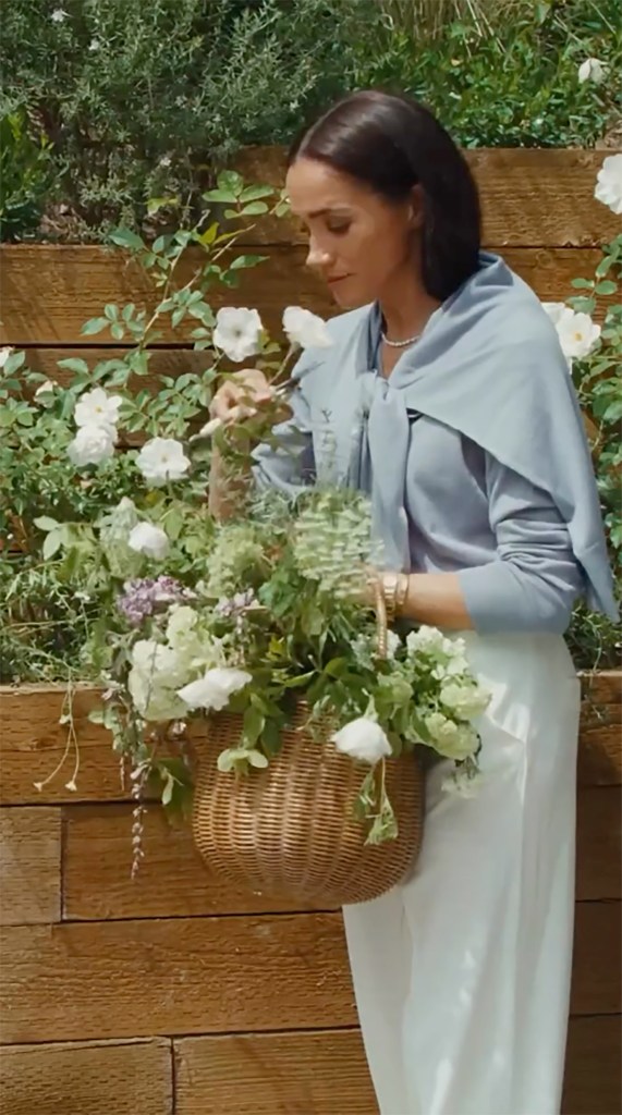 Meghan Markle arranging flowers in a wicker basket while standing next to a wooden wall.