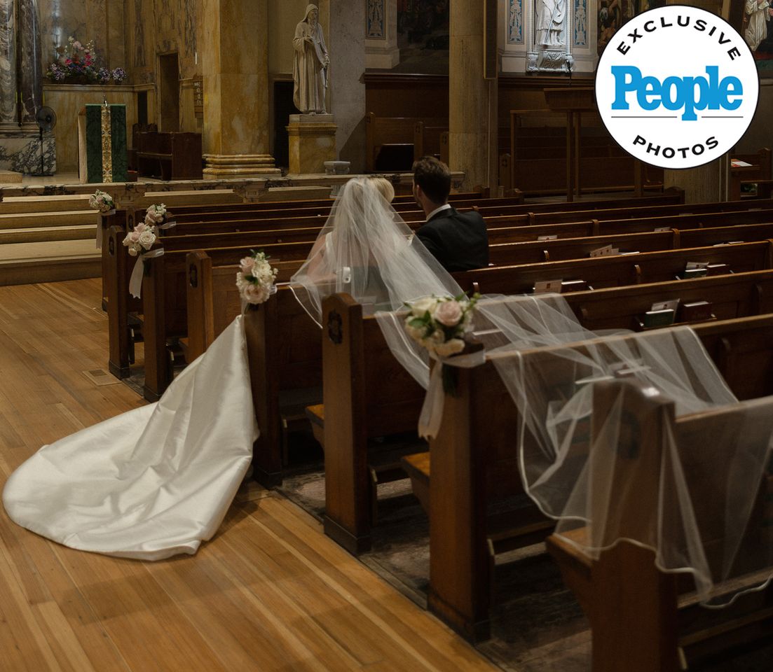 Bride and groom seated in a church with long veil and train cascading down the aisle exclusive photo branding in corner Mary Raclawski and Brandon Bussi at their wedding venueCredit: Nikki Baker Photography