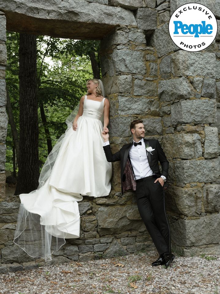 Couple dressed in wedding attire posing in a rustic outdoor setting with the bride sitting on a stone window and the groom leaning against the wall Mary Raclawski and Brandon Bussi’s wedding portraitCredit: Nikki Baker Photography