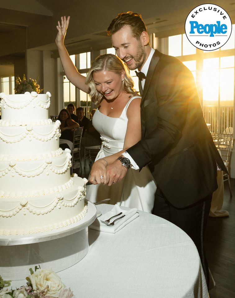 A bride and groom cutting a tiered wedding cake together with smiles set indoors with a sunset view Mary Raclawski and Brandon Bussi cutting their wedding cakeCredit: Nikki Baker Photography