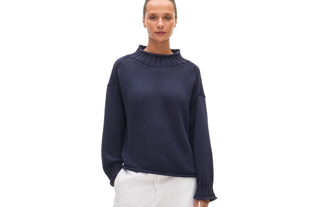 A woman wearing a dark blue, mock-neck sweater and white pants.