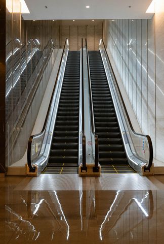 Two parallel escalators ascending in a contemporary mall, with reflective marble flooring and glass railings creating a clean, sophisticated atmosphere. A mall escalator (stock image)Credit: Getty