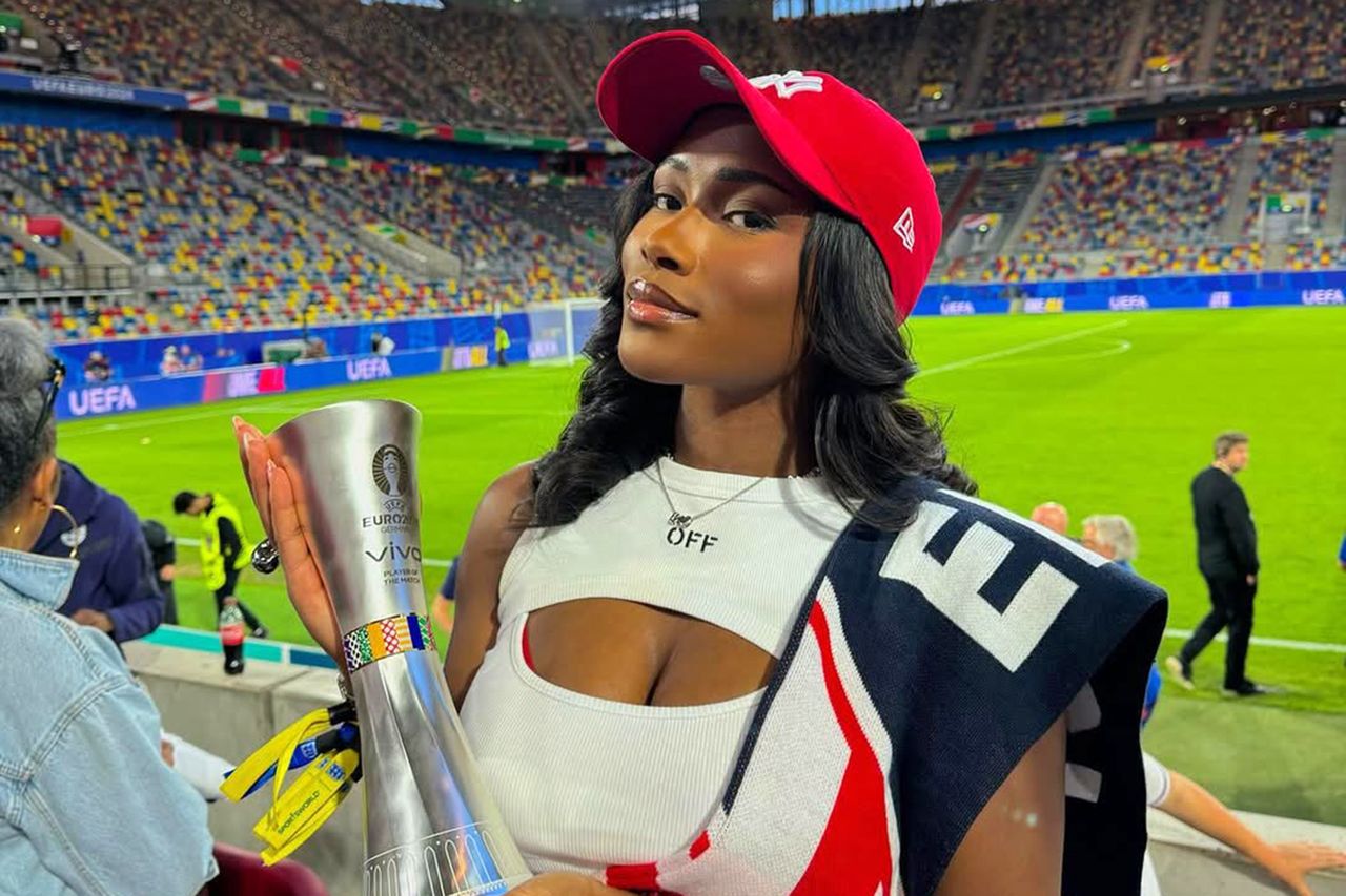 A woman in a stadium holding a trophy wearing a red cap and draped with a flag UEFA branding visible on the field in the background Tolami Benson at the 2024 Euros.Credit: tolami benson/instagram