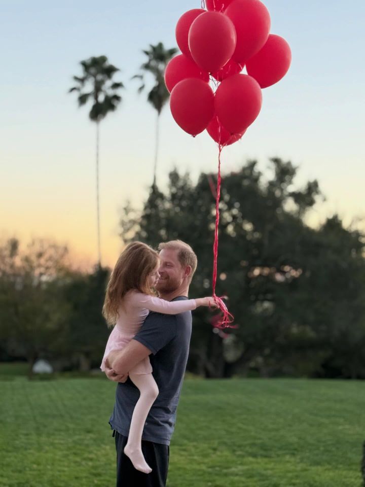 Prince Harry holds his daughter Princess Lilibet as they stand in the middle of a lawn with palm trees in the distance. Lilibet is wearing a pink ballerina ensemble and holding a bunch of red balloons. Prince Harry with Princess LilibetCredit: Meghan Markle/Instagram