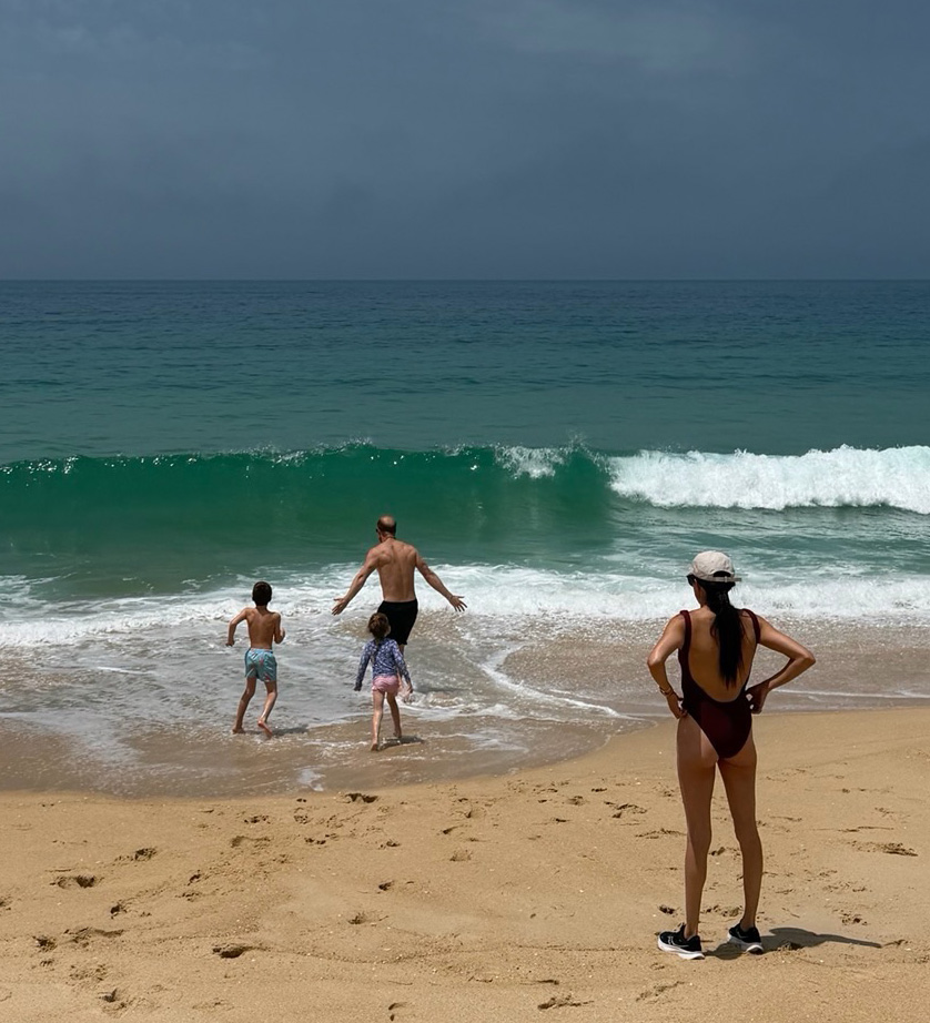 A family at the beach, a woman watches as a man and two children run into the ocean.