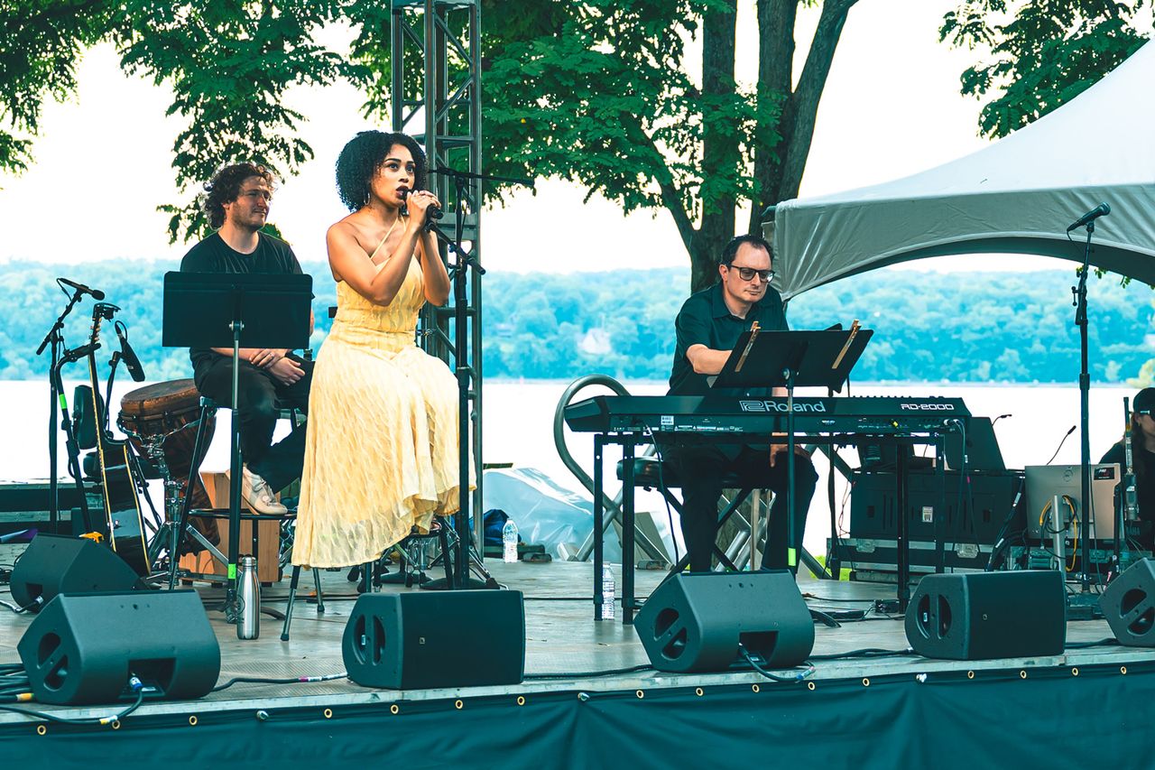 A performer singing on a stage accompanied by two musicians in an outdoor setting near a body of water Jasmine Amy Rogers sings at The Festival on Friday, Aug. 14 at Hutton Brickyards Riverfront Hotel + Venue in Kingston, N.Y.