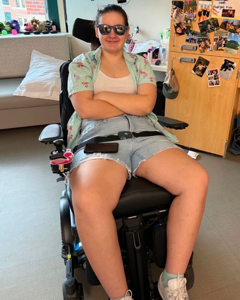 A person sitting in a wheelchair with crossed arms wearing sunglasses and casual clothes with photos and decorations visible behind Abby BuellCredit: gofundme