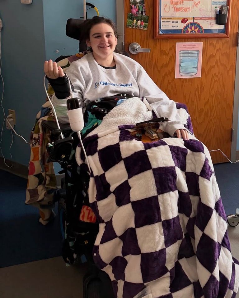 A young woman seated in a wheelchair holding a medical device with a purple checkered blanket covering her lap Abby BuellCredit: gofundme