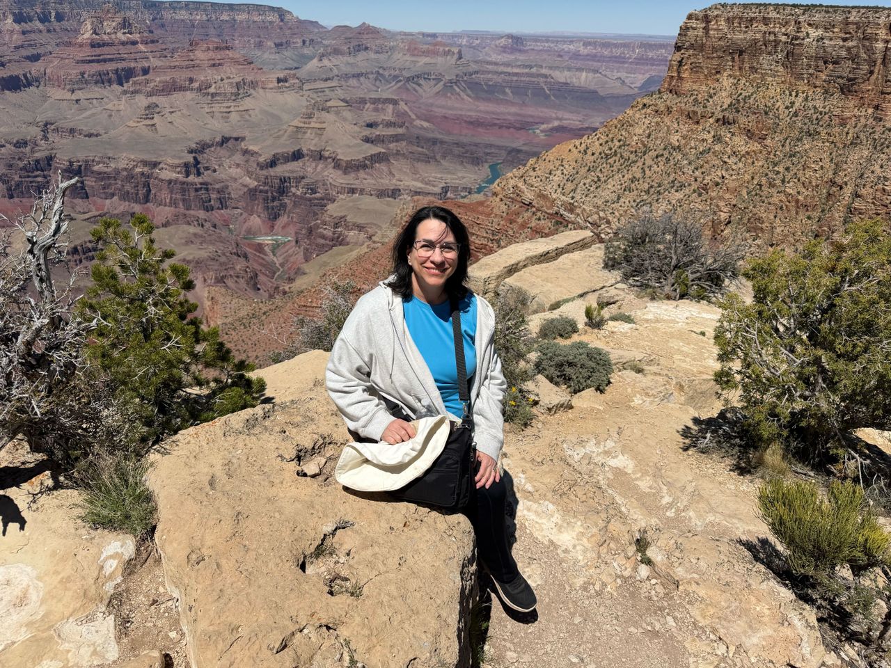 A person sitting on a rock ledge with a canyon in the background wearing casual clothing and carrying a bag Credit: People