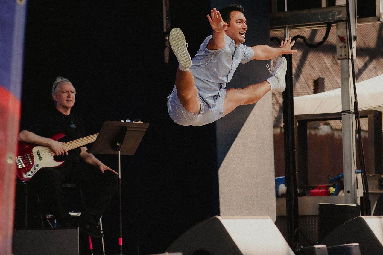 A performer in midair doing a jump with a musician playing bass seated in the background on stage Tommy Bracco shows off his moves at The Festival on Friday, Aug. 14 at Hutton Brickyards Riverfront Hotel + Venue in Kingston, N.Y.Credit: Michaelah Reynolds