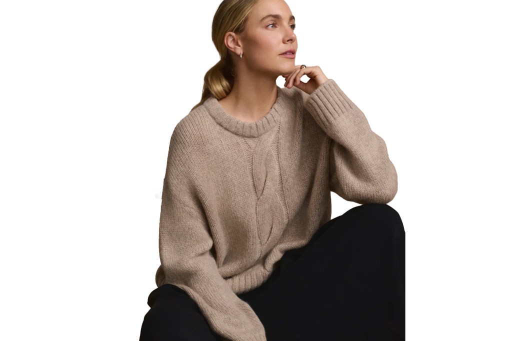 Woman with blonde hair wearing a beige knit sweater and black pants.