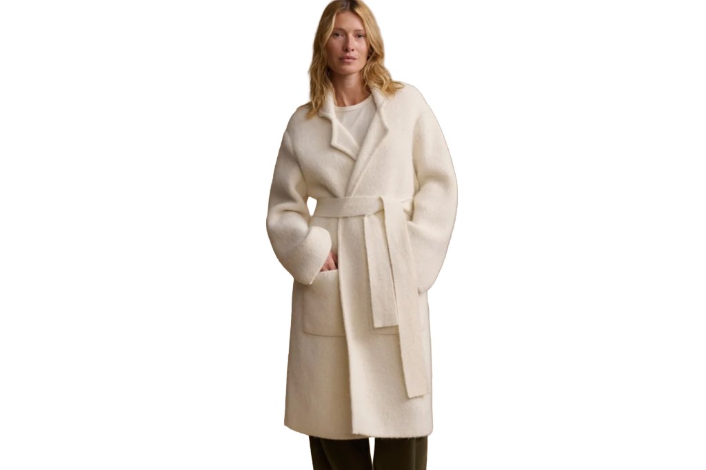 A woman models a long cream-colored coat with a belt, open collar, and pockets.