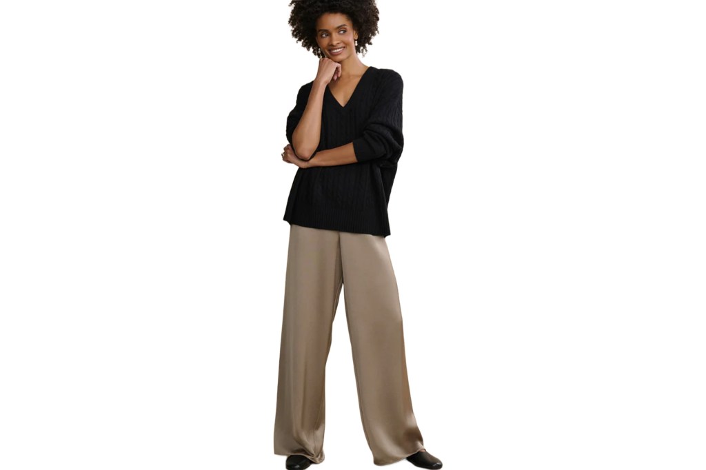 A woman in a black cable knit sweater and beige wide-leg pants smiles.