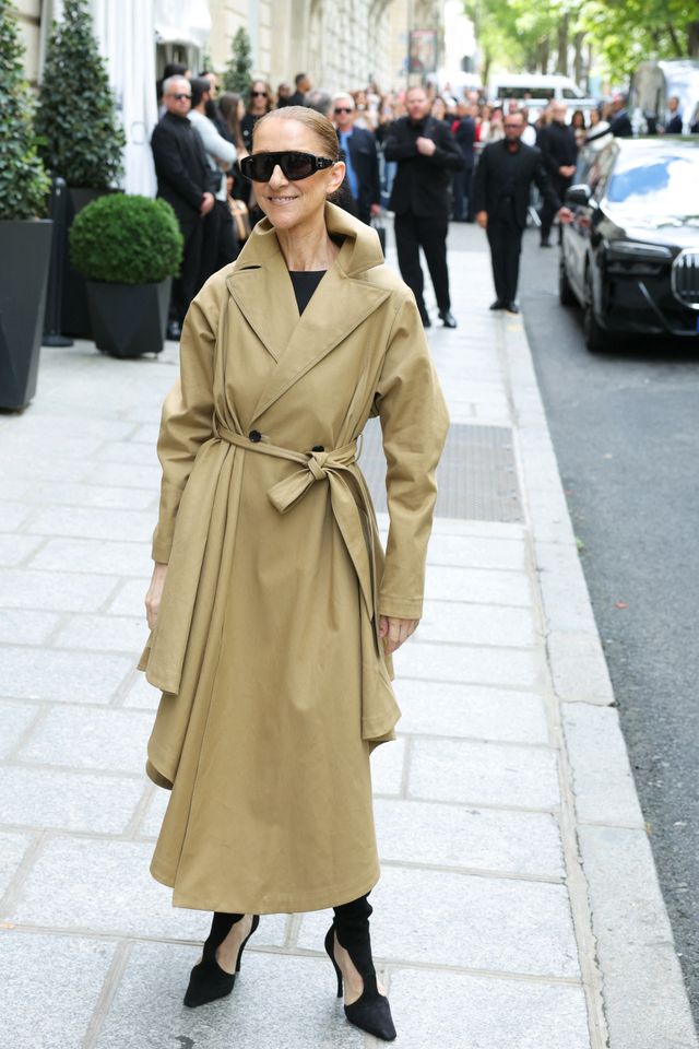 Individual wearing a trench coat and sunglasses standing outdoors with people and a car in the background Credit: Thomas SAMSON / AFP via Getty Images)